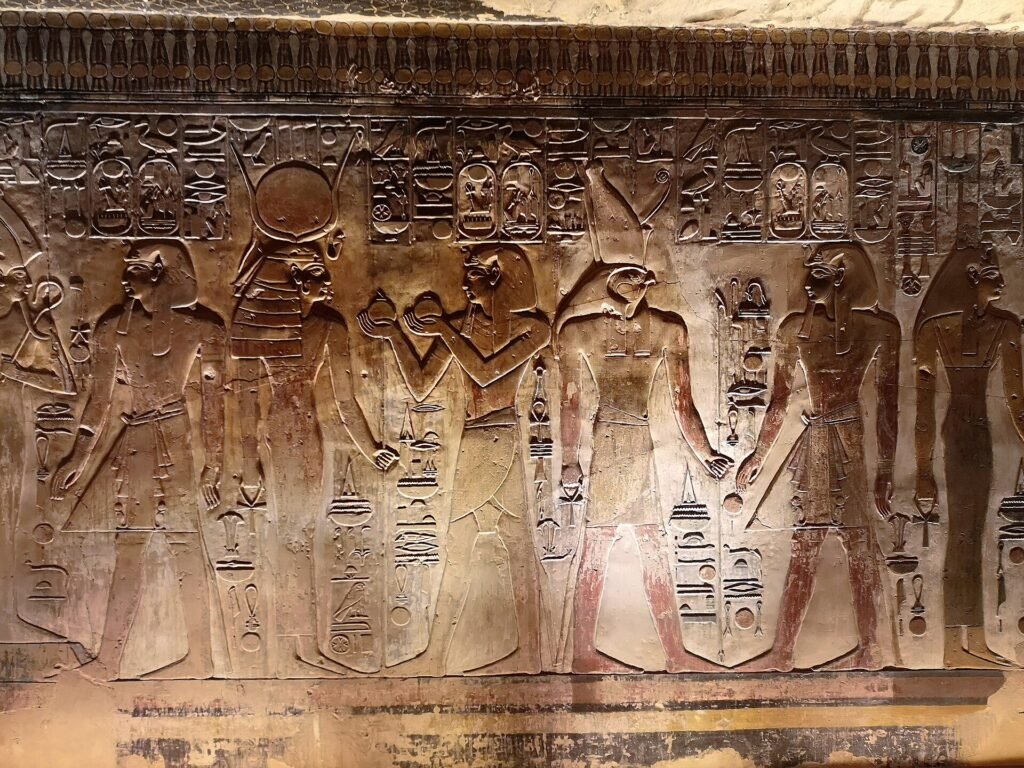 Relieve tallado de templo egipcio con procesion de dioses, una figura con cabeza de halcon estilo Horus y coronas, jeroglificos y restos de pigmento rojo sobre piedra