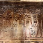 Relieve tallado de templo egipcio con procesion de dioses, una figura con cabeza de halcon estilo Horus y coronas, jeroglificos y restos de pigmento rojo sobre piedra