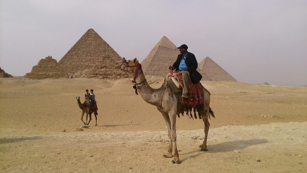 Camello ensillado con jinete frente a las tres piramides de Giza, segundo camello al fondo en el desierto, la experiencia turistica iconica en Egipto