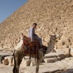 Camello blanco ensillado con manta colorida y jinete junto a los enormes bloques de piedra de la pirámide de Giza bajo cielo azul intenso