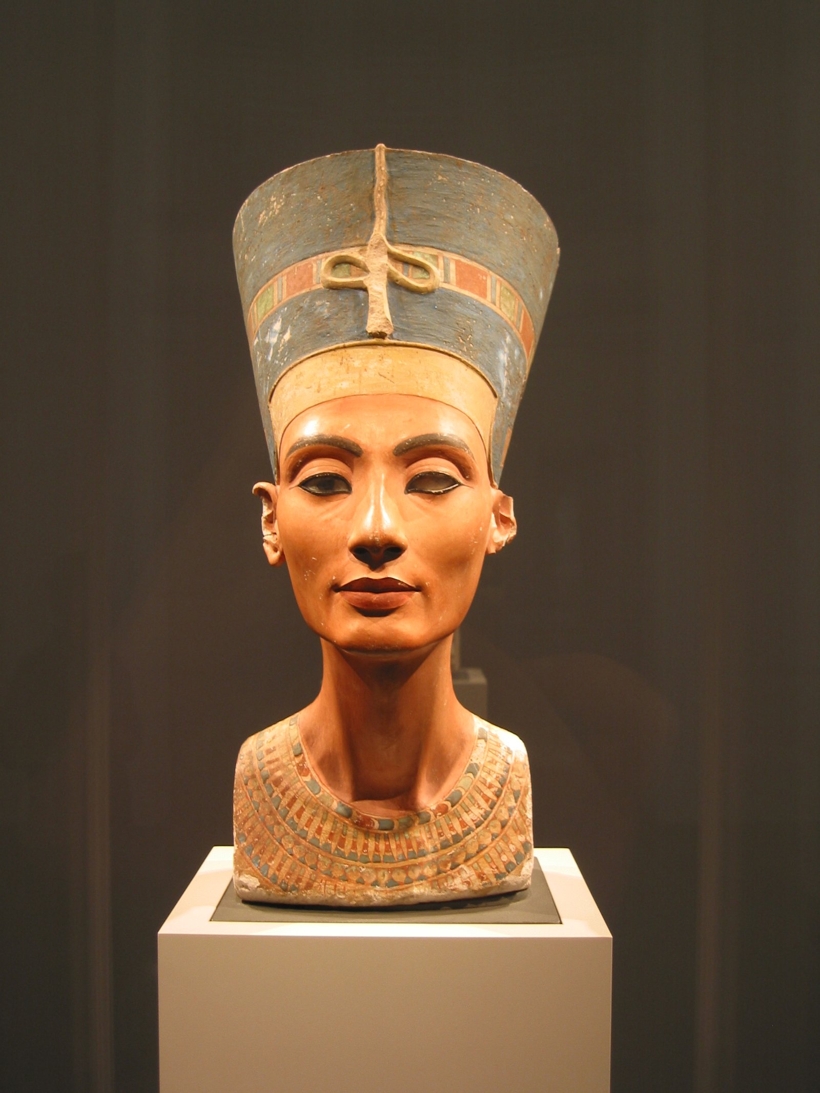 Vista frontal del famoso busto de Nefertiti sobre pedestal blanco de museo, rostro simetrico, corona azul y collar de cuentas