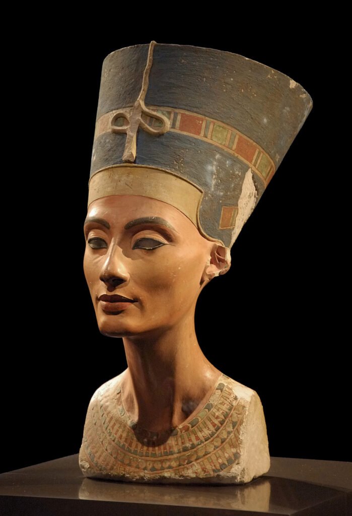 Busto policromado de la reina Nefertiti en vista de tres cuartos sobre fondo negro, con su alta corona azul y collar ancho de colores