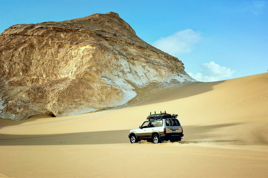 Vehiculo todoterreno de safari cruzando dunas doradas junto a una enorme formacion de tiza blanca bajo cielo azul en el Desierto Blanco de Bahariya, Egipto
