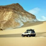 Vehiculo todoterreno de safari cruzando dunas doradas junto a una enorme formacion de tiza blanca bajo cielo azul en el Desierto Blanco de Bahariya, Egipto