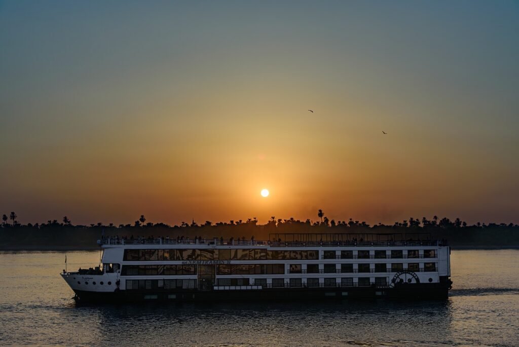Crucero por el Nilo navegando al atardecer con el sol en el horizonte sobre palmeras, reflejo dorado en el agua durante un viaje por Egipto