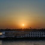 Crucero por el Nilo navegando al atardecer con el sol en el horizonte sobre palmeras, reflejo dorado en el agua durante un viaje por Egipto