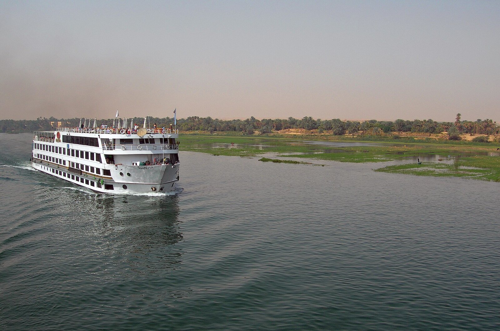 Barco de crucero por el Nilo navegando de dia entre orillas verdes y palmeras, experiencia de viaje fluvial premium en Egipto