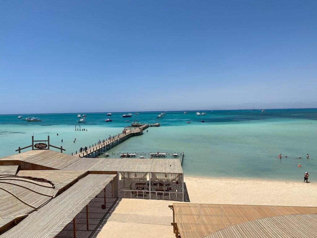 Vista elevada de Orange Bay en Hurghada con muelle de madera, barcos fondeados en aguas turquesa cristalinas del Mar Rojo y playa de arena dorada bajo cielo azul