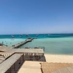 Vista elevada de Orange Bay en Hurghada con muelle de madera, barcos fondeados en aguas turquesa cristalinas del Mar Rojo y playa de arena dorada bajo cielo azul