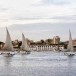 Three feluccas carrying passengers on the Nile at Luxor with a moored cruise ship and the town behind them under a soft evening sky