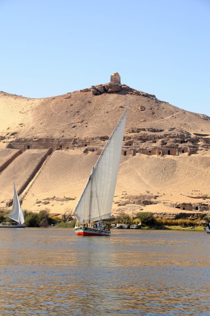 Faluca tradicional de vela blanca navegando el Nilo frente a una colina dorada del desierto en Asuan, con otra faluca al fondo bajo cielo azul despejado