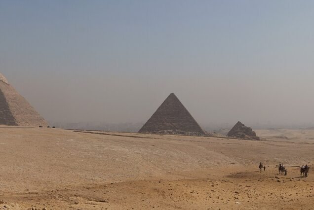 A wide panorama of all three Giza pyramids with the queens' pyramids, seen across open desert with a camel train crossing the right foreground