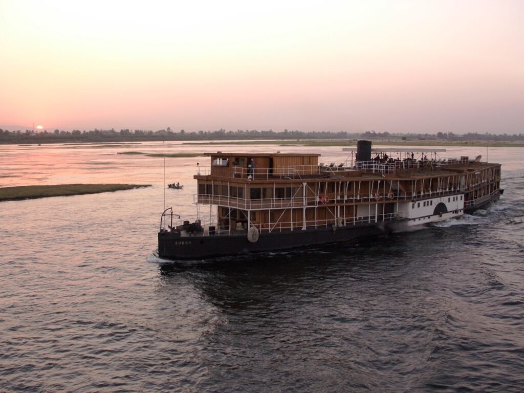 A wooden colonial style steamer on the Nile at dusk, the sun low over the far bank and passengers on the upper deck