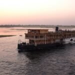 A wooden colonial style steamer on the Nile at dusk, the sun low over the far bank and passengers on the upper deck