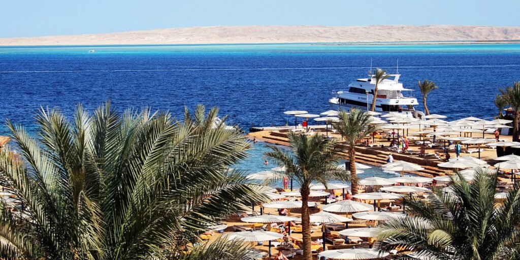 Playa de resort en Hurghada Egipto con palmeras, hileras de sombrillas blancas, un yate y aguas turquesa del Mar Rojo al fondo