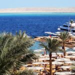 Playa de resort en Hurghada Egipto con palmeras, hileras de sombrillas blancas, un yate y aguas turquesa del Mar Rojo al fondo