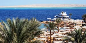 Playa de resort en Hurghada Egipto con palmeras, hileras de sombrillas blancas, un yate y aguas turquesa del Mar Rojo al fondo