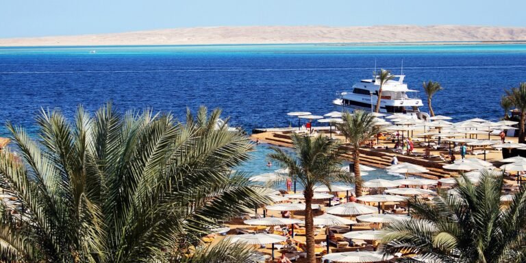 Playa de resort en Hurghada Egipto con palmeras, hileras de sombrillas blancas, un yate y aguas turquesa del Mar Rojo al fondo