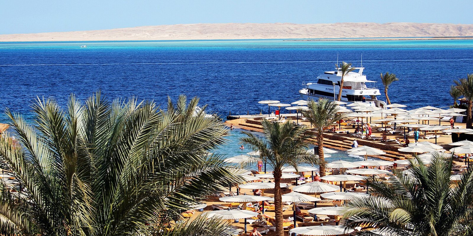 Playa de resort en Hurghada Egipto con palmeras, hileras de sombrillas blancas, un yate y aguas turquesa del Mar Rojo al fondo