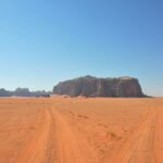 Desierto de Wadi Rum en Jordania con arena roja, huellas de neumaticos hacia un macizo de arenisca aislado y cielo azul intenso