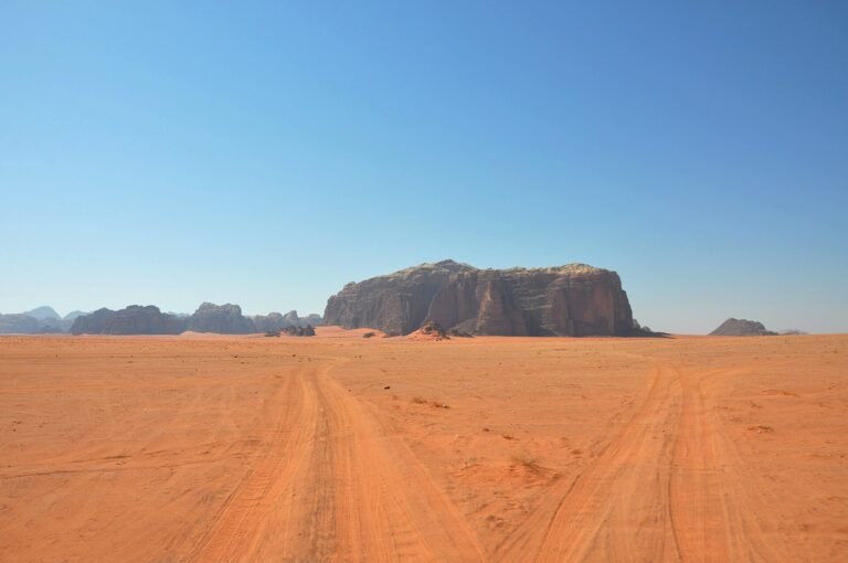 Desierto de Wadi Rum en Jordania con arena roja, huellas de neumaticos hacia un macizo de arenisca aislado y cielo azul intenso