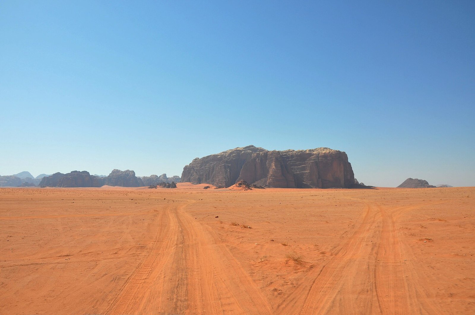 Desierto de Wadi Rum en Jordania con arena roja, huellas de neumaticos hacia un macizo de arenisca aislado y cielo azul intenso