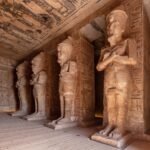 The pillared hall inside the Great Temple of Abu Simbel, with a row of colossal Osiride statues of Ramses II standing against square pillars with arms crossed over the chest, hieroglyphs and painted reliefs covering the pillars, walls and ceiling, and a wooden visitor walkway running along the floor.