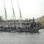 A crowded open motor launch carrying local passengers on the Nile, with a modern building and a stone embankment behind. Low resolution and flat light