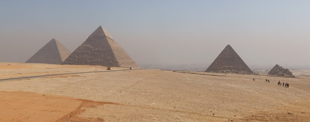 Panorama amplio de las tres pirámides de Giza sobre la arena dorada del desierto egipcio con figuras diminutas de turistas caminando a la distancia