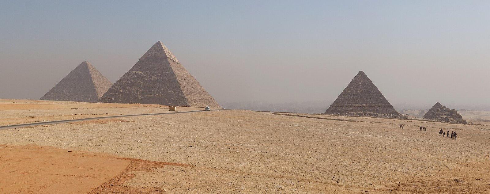 Panorama amplio de las tres piramides de Giza sobre la arena dorada del desierto egipcio con figuras diminutas de turistas caminando a la distancia