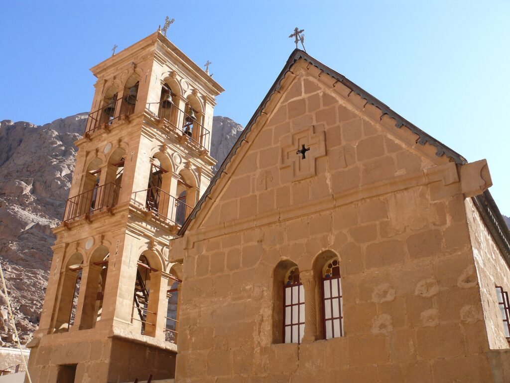 Campanario de piedra dorada del Monasterio de Santa Catalina junto a la fachada de la basilica historica con montanas del Sinai detras, Egipto