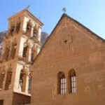 Campanario de piedra dorada del Monasterio de Santa Catalina junto a la fachada de la basilica historica con montanas del Sinai detras, Egipto