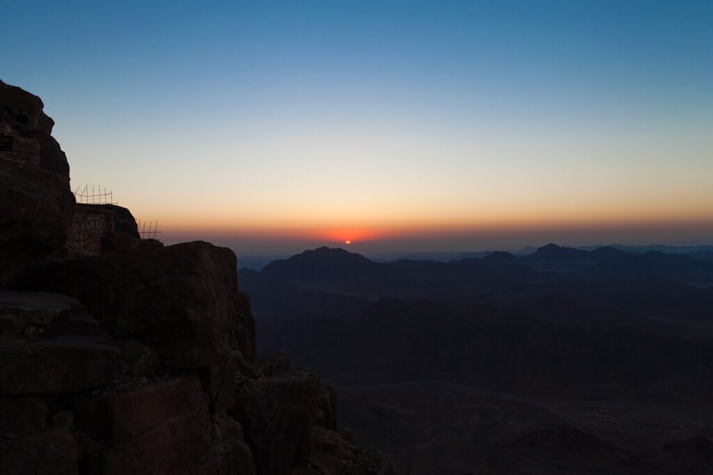 Sunrise from the summit of Mount Sinai, the sun just clearing a horizon of dark ridges with the rocky foreground in shadow. Unrelated to travel insurance