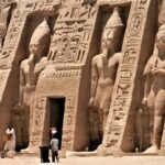 The facade of the Small Temple of Nefertari at Abu Simbel, standing colossi and deep hieroglyphic panels cut into the rock, with two people at the doorway for scale