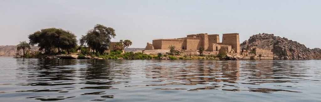 El Templo de File sobre su isla visto desde el agua del Nilo en Asuán Egipto, rodeado de palmeras y acacias con el reflejo en el río