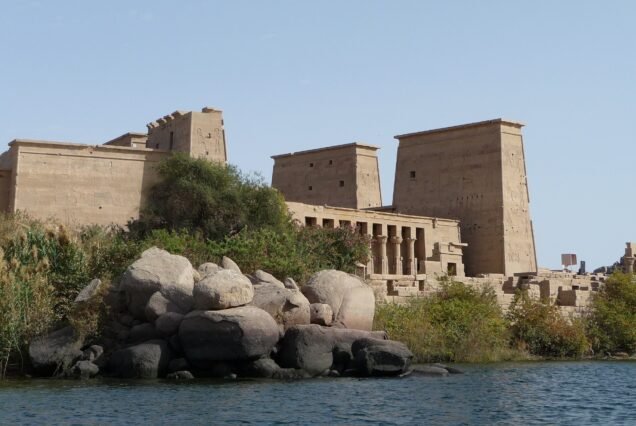 The Temple of Isis at Philae from the lake, the first pylon and colonnade above granite boulders and green scrub at the water's edge