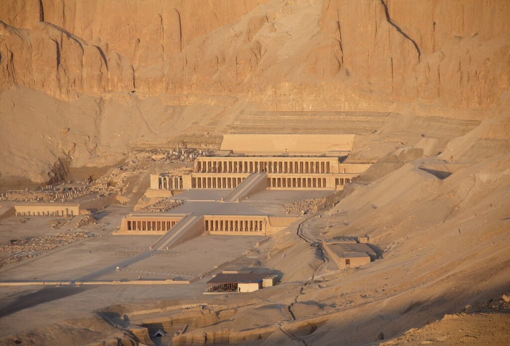 Vista elevada del Templo de Hatshepsut en Deir el-Bahari, sus terrazas escalonadas y columnatas recortadas contra los enormes acantilados dorados de Luxor al atardecer