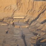 Aerial view of the terraced mortuary temple of Hatshepsut at Deir el-Bahari, its long central ramp and tiers of colonnades set against the sheer limestone cliff, with the neighbouring ruins and a visitor building on the sand around it in low golden light.