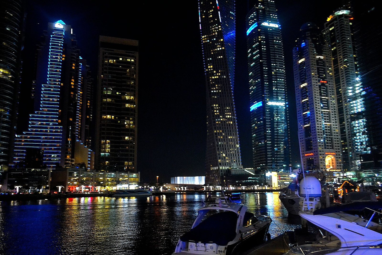 Dubai Marina iluminada de noche con torres brillantes, yates amarrados y luces reflejadas en el canal, escena nocturna de lujo en Dubai