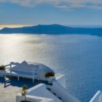 Terraza blanca de Santorini con banco y macetas sobre la caldera azul del Egeo y la isla volcanica al fondo en un dia luminoso