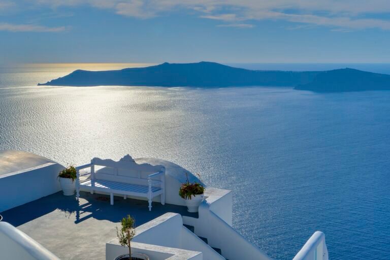 Terraza blanca de Santorini con banco y macetas sobre la caldera azul del Egeo y la isla volcanica al fondo en un dia luminoso