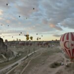 Decenas de globos aerostaticos coloridos flotando al amanecer sobre las chimeneas de hadas y formaciones rocosas de Capadocia en Turquia, con un globo de la bandera turca