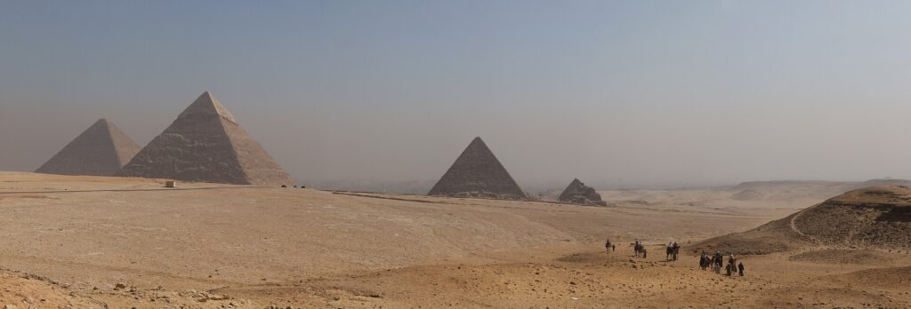 Panorama de las tres piramides de Giza en el desierto egipcio con un grupo de turistas a camello recorriendo la meseta bajo un cielo claro