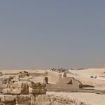 A wide panorama of the Giza plateau with the Great Sphinx on the left and the pyramids of Khafre and Khufu behind, visitors small among the ruins