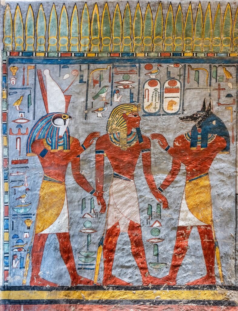 Muro pintado de una tumba del Valle de los Reyes en Luxor con Horus de cabeza de halcon, el faraon y Anubis de cabeza de chacal en rojo, azul y oro vivos