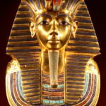 Mascara funeraria de oro de Tutankamon vista de frente, rostro dorado con tocado nemes a rayas azules y collar de gemas, tesoro del antiguo Egipto