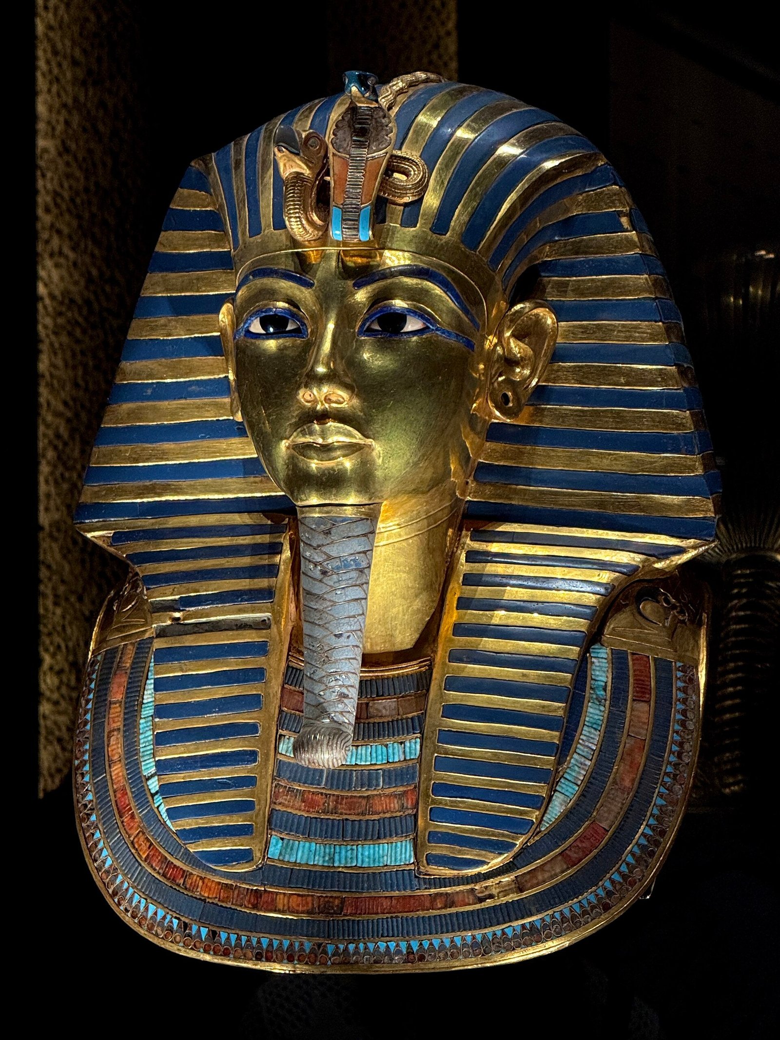 Mascara de oro de Tutankamon en vista de tres cuartos, barba trenzada y collar de lapislazuli y turquesa sobre fondo oscuro de museo