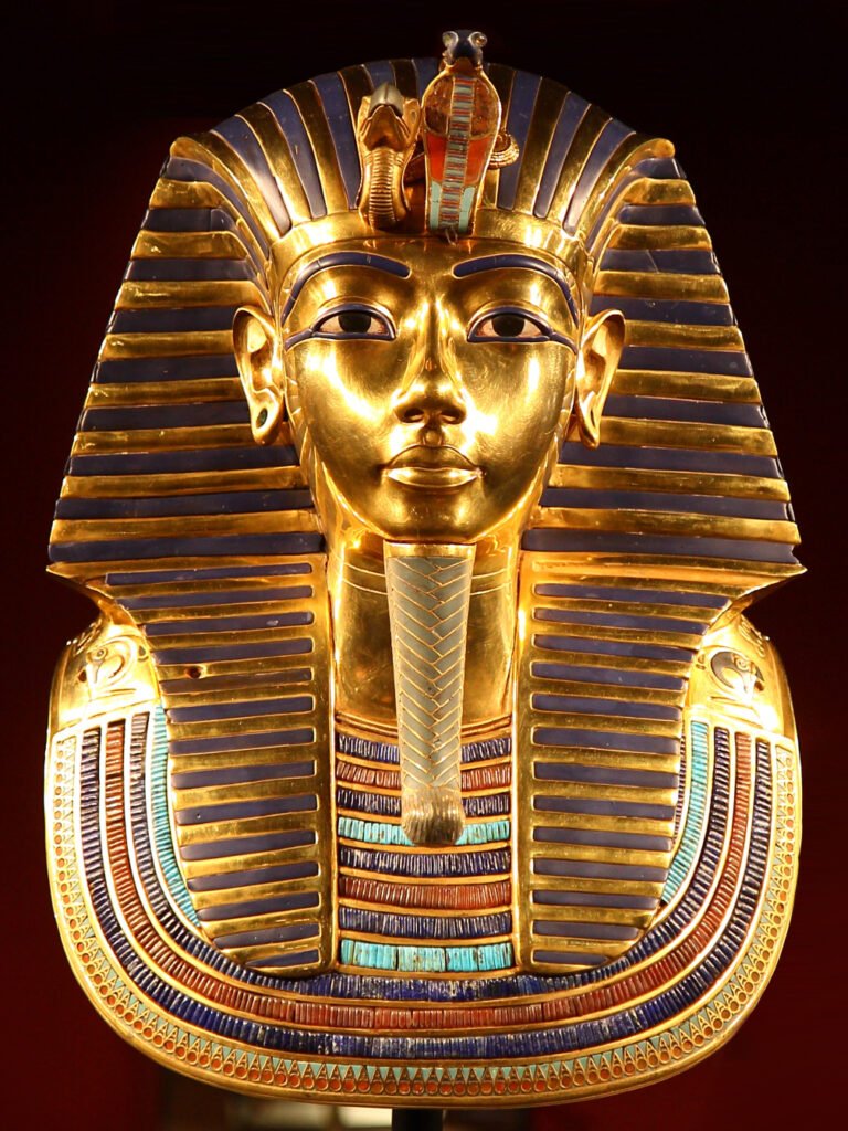 Mascara funeraria de oro de Tutankamon vista de frente, rostro dorado con tocado nemes a rayas azules y collar de gemas, tesoro del antiguo Egipto