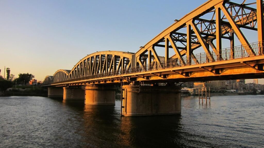 Puente de hierro sobre el rio Nilo en El Cairo bajo luz dorada del atardecer, con la estructura metalica iluminada y el horizonte urbano al fondo