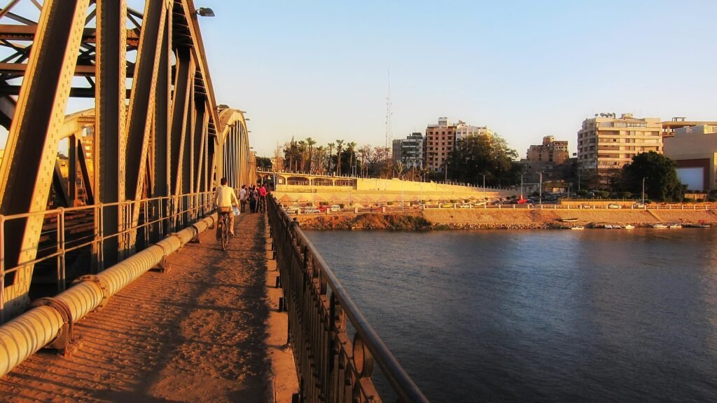 Pasarela de un puente sobre el Nilo en El Cairo al atardecer, con peatones, palmeras y edificios modernos de la ciudad iluminados por luz calida